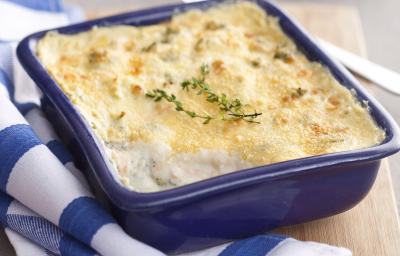 Fotografia em tons de azul em uma bancada de madeira clara, um pano branco com listras azuis, uma tábua de madeira, um recipiente quadrado azul com os legumes gratinados dentro dele. Fotografia em tons de azul em uma bancada de madeira clara, um pano branco com listras azuis, uma tábua de madeira, um recipiente quadrado azul com os legumes gratinados dentro dele.