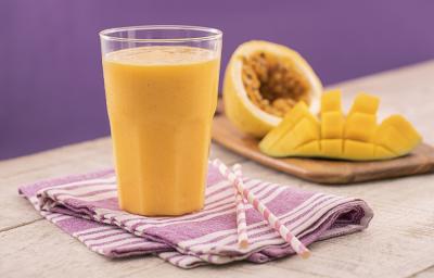 Foto em tons de roxo e amarelo da receita de smoothie de manga e maracujá servida em um copo de vidro largo sobre um pano lilás listrado com dois canudos ao lado. Ao fundo uma tábua de madeira com maracujá e manga cortados ao meio.