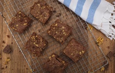 Fotografia em tons de azul em uma bancada de madeira escura, um suporte de grelha prata com alguns brownies de chocolate com nozes em cima dele e ao lado um paninho branco com listras azuis. Fotografia em tons de azul em uma bancada de madeira escura, um suporte de grelha prata com alguns brownies de chocolate com nozes em cima dele e ao lado um paninho branco com listras azuis.