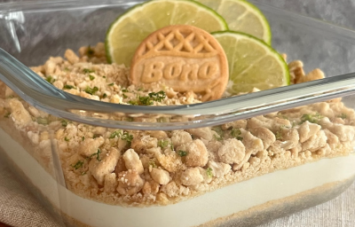 A imagem mostra um recipiente quadrado de vidro transparente, com camadas de um creme branco e biscoitos triturados. Um biscoito BONO e três rodelas de limão decoram a sobremesa.