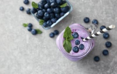 Fotografia em tons de roxo em uma bancada de madeira cinza, um copo de vidro grande com o smoothie de mirtilo dentro e um canudo branco com listras prestas e alguns mirtilos para decorar o copo. Ao lado, uma xícara com vários mirtilos. Fotografia em tons de roxo em uma bancada de madeira cinza, um copo de vidro grande com o smoothie de mirtilo dentro e um canudo branco com listras prestas e alguns mirtilos para decorar o copo. Ao lado, uma xícara com vários mirtilos.