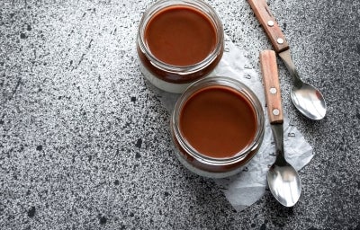 Fotografia vista de cima de dois recipientes de vidro, decorativos, com a sobremesa, finalizada com uma camada grossa de ganache. Ao lado dos recipientes tem duas colheres de sobremesa com o cabo de madeira.