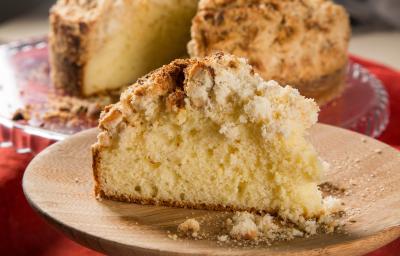 Fotografia de uma receita de cuca alemã tradicional com uma fatia grande cortada sobre um prato raso de madeira. No fundo tem a receita quase inteira sobre um apoio de vidro raso. Fotografia de uma receita de cuca alemã tradicional com uma fatia grande cortada sobre um prato raso de madeira. No fundo tem a receita quase inteira sobre um apoio de vidro raso.