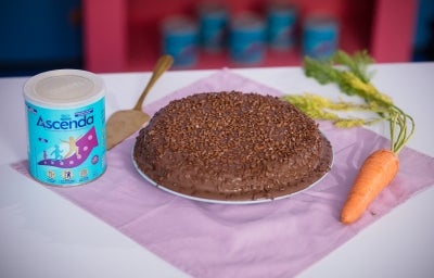 Fotografia de um bolo de cenoura com cobertura de chocolate e ascenda sobre um prato raso grande. Ao redor tem uma cenoura inteira, uma espátula e a alta de ascenda, sobre um pano roxo quadrado.