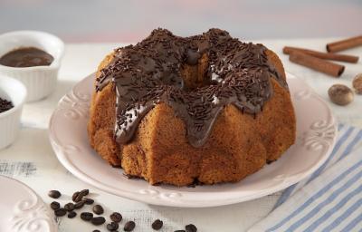Foto da receita de Bolo de Cenoura com Especiarias Coberto com Brigadeiro de Café. Observa-se um bolo inteiro em cima de um prato de porcelana branco com brigadeiro por cima e granulado. Grãos de café, canela em pau e o brigadeiro decoram a foto.