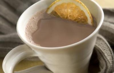Fotografia em tons de cinza em uma bancada de madeira clara, um pano cinza escuro, uma xícara branca com o chocolate quente com uma rodela de laranja dentro.
