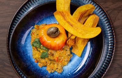 Fotografia em tons de marrom com um prato preto ao centro. Em cima do prato existe uma receita de ceviche de caju com castanhas e chips de banana da terra Fotografia em tons de marrom com um prato preto ao centro. Em cima do prato existe uma receita de ceviche de caju com castanhas e chips de banana da terra