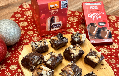 Foto da receita de palha italiana de mini panettone brasil cacau, servida em pedaços pequenos sobre uma tábua de madeira, em uma bancada decorada com itens natalinos e produtos da marca brasil cacau ao redor Foto da receita de palha italiana de mini panettone brasil cacau, servida em pedaços pequenos sobre uma tábua de madeira, em uma bancada decorada com itens natalinos e produtos da marca brasil cacau ao redor