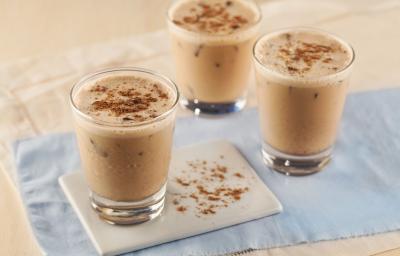 Fotografia em tons de azul em uma bancada de madeira clara com um paninho azul e três copinhos de vidro com a bebida de doce de leite, Leite Moça e Nescau.