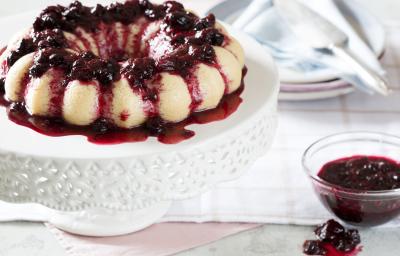 foto em tons de branco e vinho tirada de uma mesa branca, contém um suporte para servir sobremesas branco com o Brigadeirão Branco com Calda de Frutas Vermelhas. Ao lado um pote transparente redondo com a calda de frutas vermelhas. foto em tons de branco e vinho tirada de uma mesa branca, contém um suporte para servir sobremesas branco com o Brigadeirão Branco com Calda de Frutas Vermelhas. Ao lado um pote transparente redondo com a calda de frutas vermelhas.