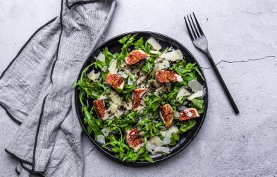 Fotografia vista de cima de uma salada de folhas, figo, requeijão e molho de maracujá e com vinagre balsâmico. A salada está em um prato raso preto, e ao lado, um garfo preto, sobre uma mesa com tons de cimento.