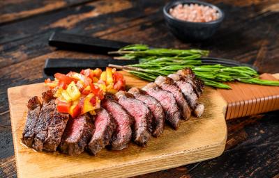Fotografia em tons de marrom em uma bancada de madeira preta. Ao centro, uma tábua de madeira contendo a picanha fatiada ao fundo há duas facas, temperos e um recipiente contendo sal rosa.