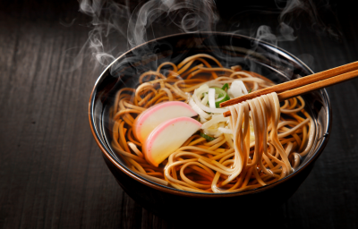 Fotografia mostra um macarrão japonês sendo segurado por hashi, ao fundo um bowl escuro com macarrão dentro.