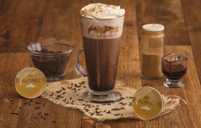 Fotografia em tons de marrom em uma bancada de madeira com um copo grande de vidro com a bebida de Alpino, chocolate quente. Ao fundo, cápsulas de alpino da Dolce Gusto, um vidrinho de canela, um copinho com raspas de chocolate.