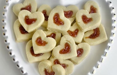 Fotografia em tons claros com vários biscoitos em formato de coração ao centro. No centro do cada coração, existe uma pequena quantidade de goiabada derretida