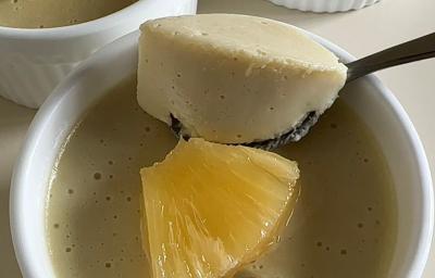 Fotografia em tons claros de três ramekins brancos com mousse de abacaxi com um pedaço da fruta decorando por cima. O ramekin no centro da foto tem uma colher segurando um pouco da mousse.