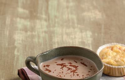 Fotografia em tons de marrom em uma bancada de madeira com um pano xadrez roxo com uma caneca grande com o chocolate quente feito com bombom de Alpino.