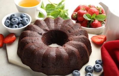 Bolo de chocolate em forma de bundt, com superfície brilhante e densa, servido em uma tábua clara. Ao redor, chá, folhas de hortelã e morangos frescos.