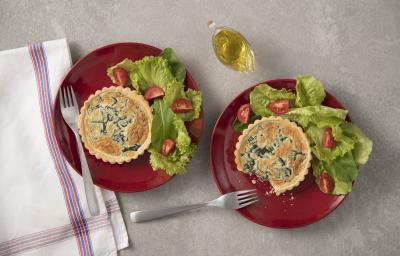 Fotografia em tons de cinza, branco, vermelho e verde de uma bancada cinza vista de cima. Contém dois pratos redondos vermelhos ambos com uma tortinha e ao lado folhas de alface com fatias de tomate e garfos para servir. Ao lado temos um pano branco. Fotografia em tons de cinza, branco, vermelho e verde de uma bancada cinza vista de cima. Contém dois pratos redondos vermelhos ambos com uma tortinha e ao lado folhas de alface com fatias de tomate e garfos para servir. Ao lado temos um pano branco.