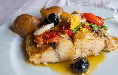 Em cima de uma mesa, prato com porção de bacalhau com azeite e tomate, pimentão, batatas e azeitonas. Em cima de uma mesa, prato com porção de bacalhau com azeite e tomate, pimentão, batatas e azeitonas.