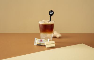 Foto da receita de Espresso Tônica. Observa-se um copo alto com gelo e duas fases aparentes: metade tônica e metade café. O cenário é bege e está decorado com gengibre e servido com um KIT KAT Branco.