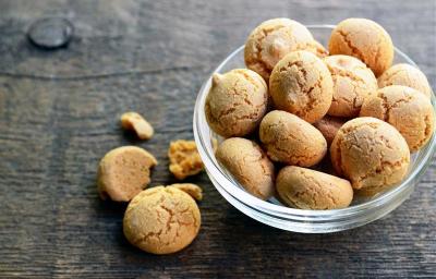Fotografia de um recipiente de vidro médio fundo com biscoitos de amêndoas tipo amaretti, ao lado tem um e um pedaço sobre uma mesa de madeira.