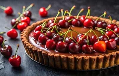 Fotografia em tons escuros de uma torta de chocolate e amarenas sobre uma mesa preta. Ao redor tem cerejas inteiras e também sobre a torta. Fotografia em tons escuros de uma torta de chocolate e amarenas sobre uma mesa preta. Ao redor tem cerejas inteiras e também sobre a torta.