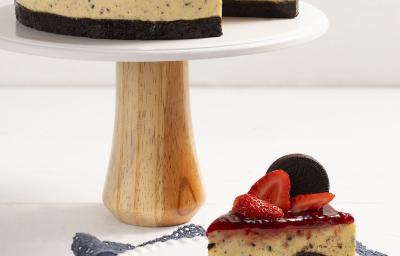 Fotografia em tons de branco em uma bancada de madeira em tons de branco com um pano marrom. No centro, um prato branco contendo uma fatia do cheesecake com um garfo ao lado e atrás está o restante do cheesecake. Fotografia em tons de branco em uma bancada de madeira em tons de branco com um pano marrom. No centro, um prato branco contendo uma fatia do cheesecake com um garfo ao lado e atrás está o restante do cheesecake.