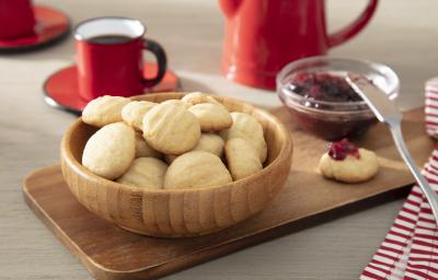 Foto da receita de biscoito de nata. Observa-se os biscoitinhos dentro de um recipiente fundo e redondo de madeira. Ao lado, um dos biscoitinhos está com geléia de morango e atrás tem uma xícara com café.