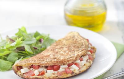 Fotografia em tons de verde em uma mesa de madeira cinza com um pano verde, um prato branco raso redondo e a omelete integral recheada com queijo branco, tomate e ervas, ao lado salada para acompanhar. Ao fundo, um potinho com azeite. Fotografia em tons de verde em uma mesa de madeira cinza com um pano verde, um prato branco raso redondo e a omelete integral recheada com queijo branco, tomate e ervas, ao lado salada para acompanhar. Ao fundo, um potinho com azeite.