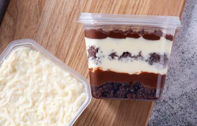 Fotografia em tons de marrom com um pote de bolo ao centro. Dentro desse pote existe camadas de um creme de cor branca e camadas de um creme de cor chocolate. Em cima do bolo existe coco ralado Fotografia em tons de marrom com um pote de bolo ao centro. Dentro desse pote existe camadas de um creme de cor branca e camadas de um creme de cor chocolate. Em cima do bolo existe coco ralado