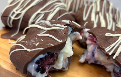Fotografia de uma tábua de madeira clara sobre uma mesa cinza claro. Sobre a tábua tem alguns bombons coobertos com chocolate meio amargo e decorados com chocolate branco, o recheio é um creme de ninho com frutas vermelhas, um deles está aberto. Fotografia de uma tábua de madeira clara sobre uma mesa cinza claro. Sobre a tábua tem alguns bombons coobertos com chocolate meio amargo e decorados com chocolate branco, o recheio é um creme de ninho com frutas vermelhas, um deles está aberto.