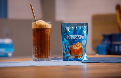 Copo alto com bebida escura e espuma ao lado de um sachê azul de Nescafé Ice sobre uma mesa.
