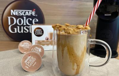 Foto em tons de marrom e bege com uma caneca de vidro situada ao centro. Dentro da caneca existe uma preparação de café da cor caramelo decorada com pedaços de biscoito por cima do copo. Ao fundo existe uma caixa de cápsulas de café da marca Dolce Gusto