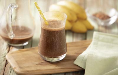 Fotografia em tons de marrom e amarelo de uma bancada de madeira com uma tábua, um paninho verde e um copo com a bebida dentro e um canudo de papel amarelo. Ao fundo uma jarra com a bebida, bananas e cacau. Fotografia em tons de marrom e amarelo de uma bancada de madeira com uma tábua, um paninho verde e um copo com a bebida dentro e um canudo de papel amarelo. Ao fundo uma jarra com a bebida, bananas e cacau.