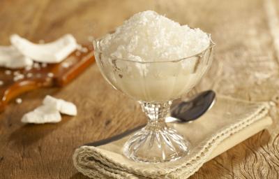 Fotografia em tons de marrom de uma bancada marrom, sobre ela um paninho beje, um taça com raspadinha e uma colher. Ao fundo uma tábua com pedaços de coco. Fotografia em tons de marrom de uma bancada marrom, sobre ela um paninho beje, um taça com raspadinha e uma colher. Ao fundo uma tábua com pedaços de coco.