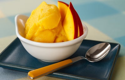 Imagem de um bowl branco com sorvete de manga e fatias de manga e uma colher ao lado.