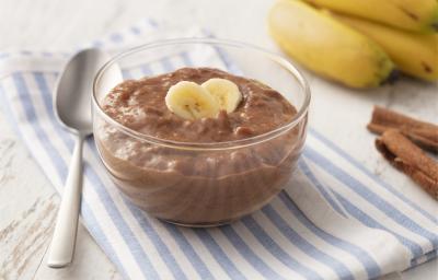 Fotografia em tons de azul em uma bancada de madeira clara com um paninho listrado azul e em cima um potinho de vidro com o doce de banana e cacau. Ao lado, algumas bananas, uma colher e canela em pau. Fotografia em tons de azul em uma bancada de madeira clara com um paninho listrado azul e em cima um potinho de vidro com o doce de banana e cacau. Ao lado, algumas bananas, uma colher e canela em pau.