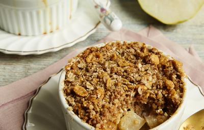 fotografia em tons de cinza e marrom tirada de uma bancada cinza, contém dois pratos redondos e branco, e ambos contém um pote redondo branco com o Crumble. Ao lado direito contém duas fatias de pera. fotografia em tons de cinza e marrom tirada de uma bancada cinza, contém dois pratos redondos e branco, e ambos contém um pote redondo branco com o Crumble. Ao lado direito contém duas fatias de pera.