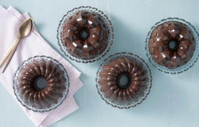 Fotografia em tons de chocolate, azul e rosa, com quatro porções de pudim de chocolate em pratinhos de cristal, um deles sobre guardanapo rosa tendo ao lado colheres de sobremesa, tudo sobre bancada azul.