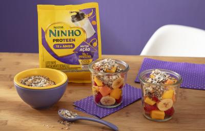 Fotografia de dois copos de vidro com frutas e granola sobre um paninho roxos com bolinhas brancas, sobre uma mesa de madeira. Ao lado dos copos, tem um recipiente amarelo e roxo com a granola, e tem uma embalagem de ninho proteen. Fotografia de dois copos de vidro com frutas e granola sobre um paninho roxos com bolinhas brancas, sobre uma mesa de madeira. Ao lado dos copos, tem um recipiente amarelo e roxo com a granola, e tem uma embalagem de ninho proteen.