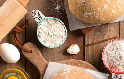 Fotografia em tons de branco e marrom, com massas para macarrão sobre papel manteiga e peças de madeira, no entorno potinho com ovo. ovo inteiro, potinhos com farinha de trigo, colher com sal, rolo de abrir massa, tudo sobre bancada de madeira.