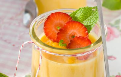 Fotografia de dois recipientes de vidro decorativos com uma mousse de laranja, no recipiente da frente, em cima da mousse, há três fatias de morangos com hortelã ao lado. Ao fundo, no recipiente há apenas a mousse de laranja. Fotografia de dois recipientes de vidro decorativos com uma mousse de laranja, no recipiente da frente, em cima da mousse, há três fatias de morangos com hortelã ao lado. Ao fundo, no recipiente há apenas a mousse de laranja.