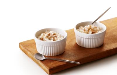 Foto da receita de Arroz Doce Vegano. Observa-se uma tábua de madeira com dois potinhos brancos e pequenos de arroz doce em cima. Foto da receita de Arroz Doce Vegano. Observa-se uma tábua de madeira com dois potinhos brancos e pequenos de arroz doce em cima.