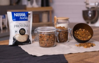 Foto da receita de granola proteica com aveia servida em dois potes herméticos de vidro sobre um pano neutro em cima de uma mesa de madeira com uma embalagem de aveia proteína ao lado