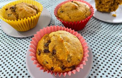 Fotografia de três forminhas de cupcake com bolo de cenoura e uva passa, sendo duas de tom rosa e uma amarela. Duas delas estão sobre um miniprato cinza raso, os quais estão sobre uma toalha de mesa de tom azul claro com bolinhas pretas.