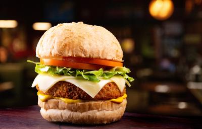 Fotografia de um hamburguer com tomate, alface, queijo, hambúrguer empanado e um molho amarelo situado ao centro de uma foto em tons escuros