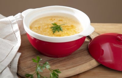 Fotografia de um recipiente pequeno e fundo, vermelho e branco, sobre uma tábua de madeira pequena ao lado da tampa e de uma pano branco. Dentro do recipiente tem o creme de cenoura e mandioquinha.
