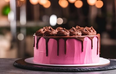 Fotografia de um bolo redondo coberto com um vibrante glacê, adornado com ganache de chocolate brilhante que escorre pelas laterais. O fundo desfocado com luzes quentes cria uma atmosfera acolhedora.