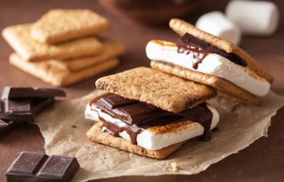 Fotografia de duas peças de s'mores sobre um papel manteiga. Ao fundo tem alguns pedaços de chocolate e alguns biscoitos empilhados, sobre uma mesa de madeira.